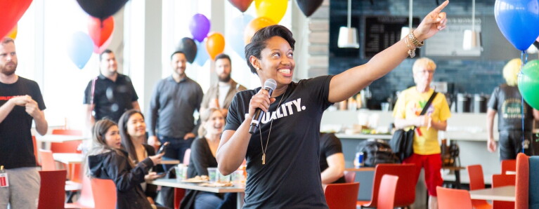 A Black woman wearing a shirt that says "Equality" speaks into a microphone while gesturing off camera. She is speaking to a brightly decorated room full of people.