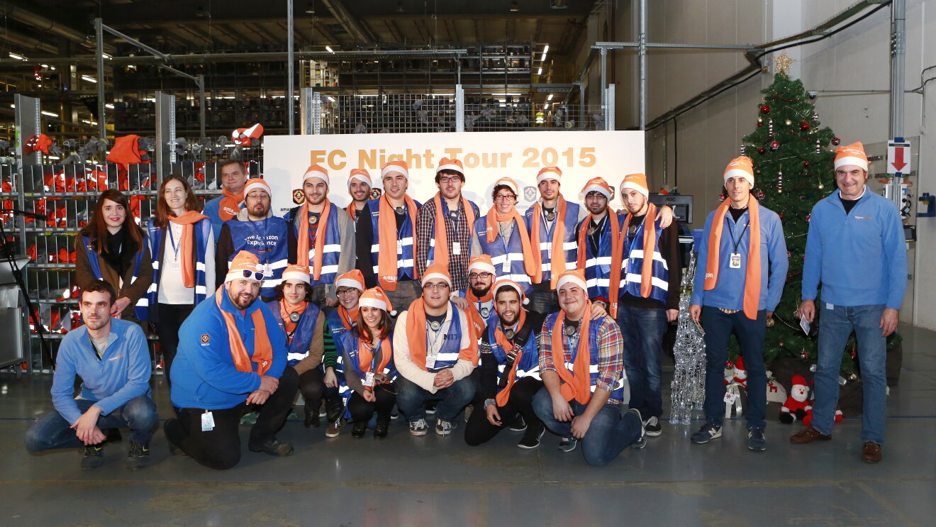 Foto de grupo de 23 personas posando delante de un cartel FC Night Tour 2015. Algunos de ellos lucen una bufanda de color naranja y un chaleco de color azul. 