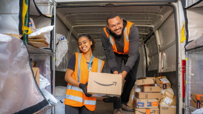 No fundo de um furgão, um homem e uma mulher da equipe de entrega carregam uma caixa. Eles sorriem