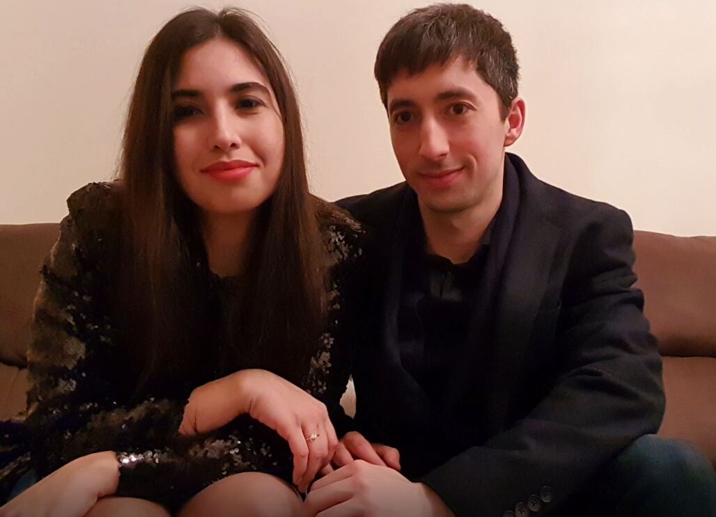 A brunette woman and brunette man sit together on a sofa, slight smiles on their faces. 