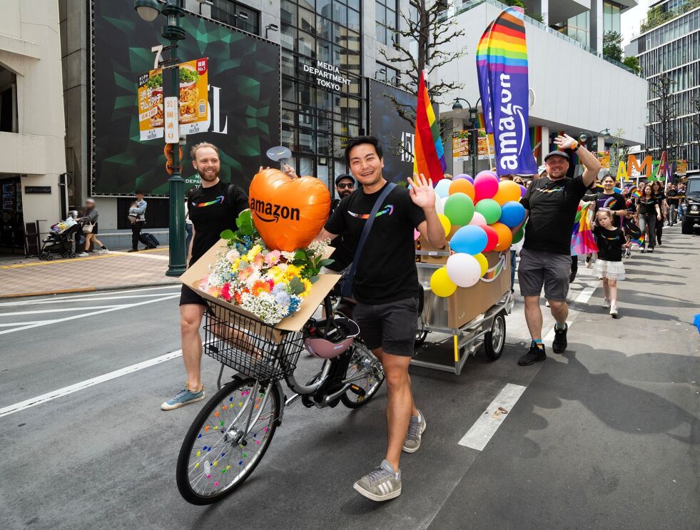 風船や花などで装飾されたリヤカー付き自転車を押している3人の男性が手を振っている。彼らの後ろにパレードの列が続いている