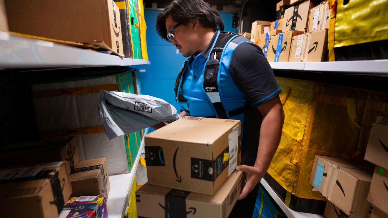青い制服とスマートグラスを着用した配送員が、倉庫内の棚の間に立って荷物を取り出している。棚にはAmazonロゴのついた段ボール箱が多数。