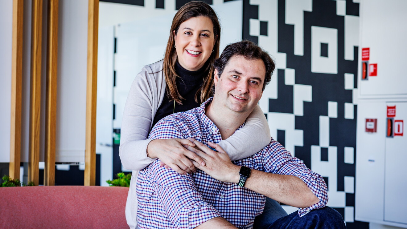 Ane y Pablo posando en las oficinas corporativas en Madrid para la sesión de fotos del post que tenemos entre manos. Los dos están sentados en un sofá de color rosa. Él delante sentado y ella detrás, de rodillas agarrando a si maridopor el cuello. Pablo le agarra también las manos. Él lleva una camisa de cuadros de color rojo y azul y ella lleva el pelo suelto, un jersey de cuello alto de color negro y por encima una chaqueta de punto. De fondo se ven unas plantas y una pared estampada de color negro y blanco. 