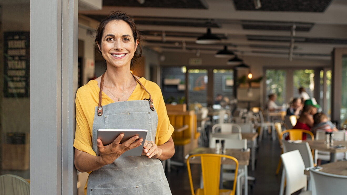A woman holding a tablet and wearing an apron leans against a post and smiles at the camera. Behind her people are seated at tables in a cafe or restaurant. 