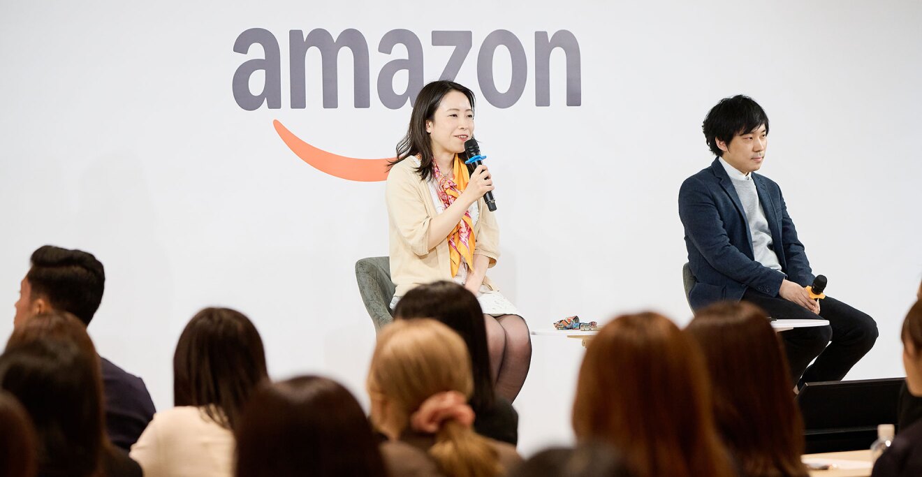 Amazonのロゴが大きく書かれた壁を背に、聴衆の前で座って話をする2人の人物。2院とも手にマイクを持っている。手前に聴衆の後頭部が並ぶ。