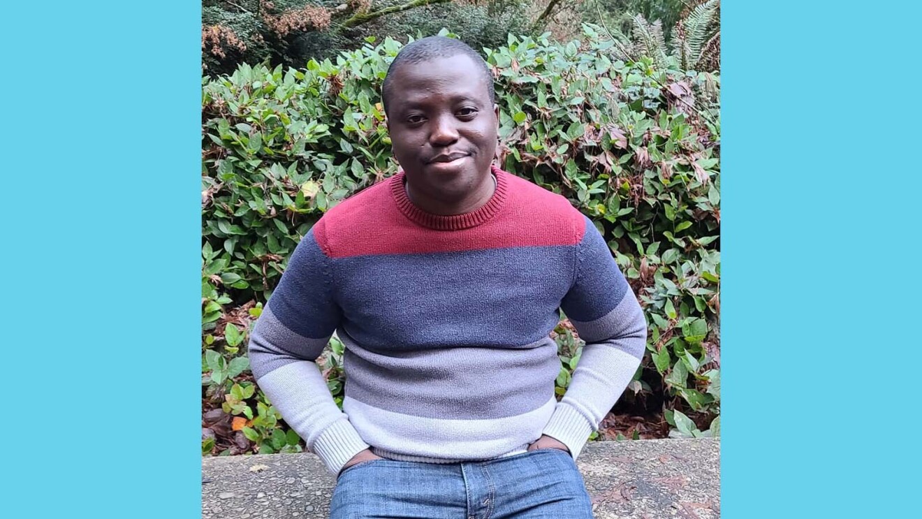 Olabanji smiling and casually sitting on a bench in front of a bush and trees.