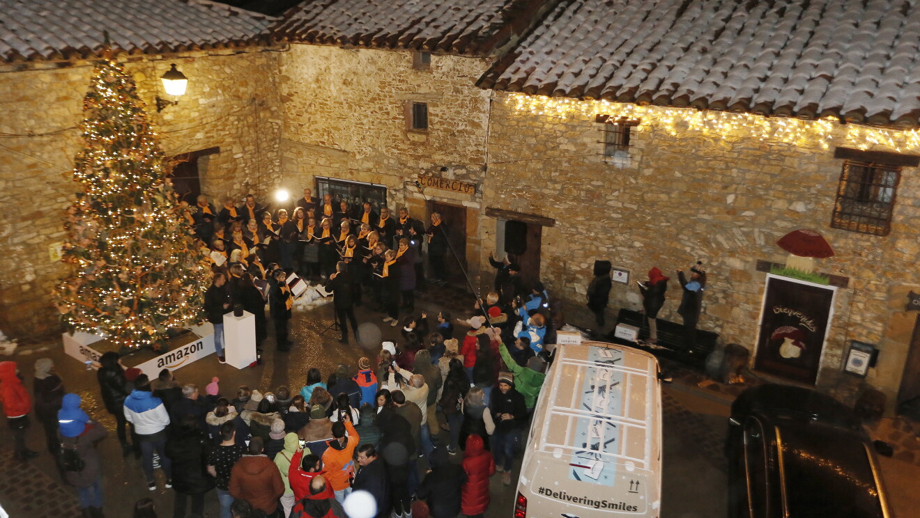 En la plaza principal de Valdelinares rodeada de tres casa de piedra con nieve en el tejado se encuentra: un árbol de Navidad decorado con luces y detrás un coor de 40 cantantes vestidos de negro y con una bufanda de color amarillo. Rodeando el árbil y el coro, los vecinos del pueblo. 