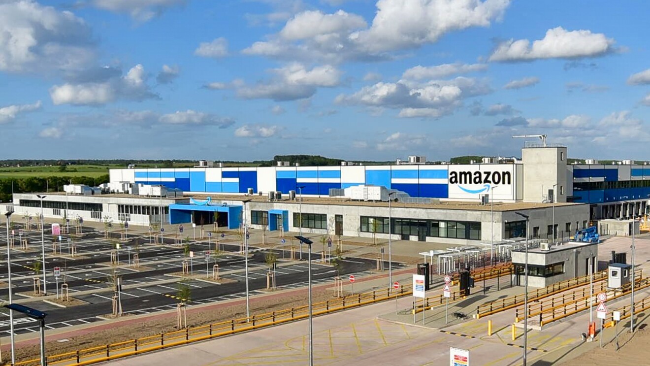 Ein weiß-blaues langgezogenes Gebäude mit Flachdach. Auf der Frontseite sieht man ein großes Amazon Logo mit blauem Smile-Pfeil. Vor dem Gebäude wurde ein großzügiger Mitarbeiterparkplatz mit Baum-Begrünungen angelegt.