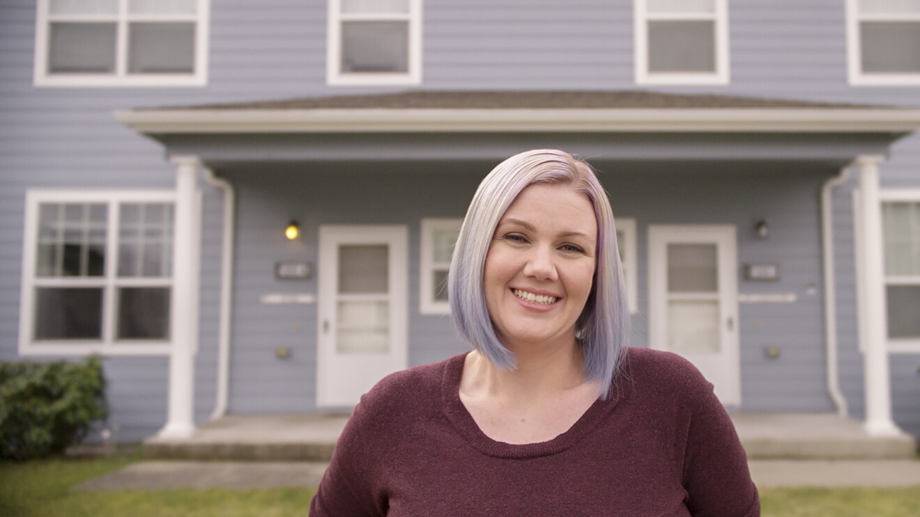 A woman stands in front of her home, smiling at the camera.