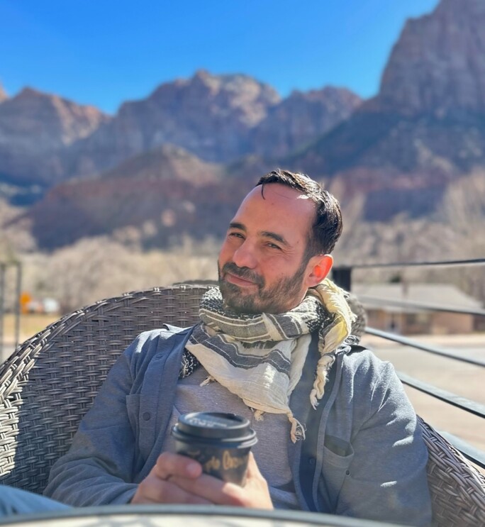 Chief Meteorologist Victor Rodriguez holds a coffee in his hands as he sits in a chair near the base of mountains.