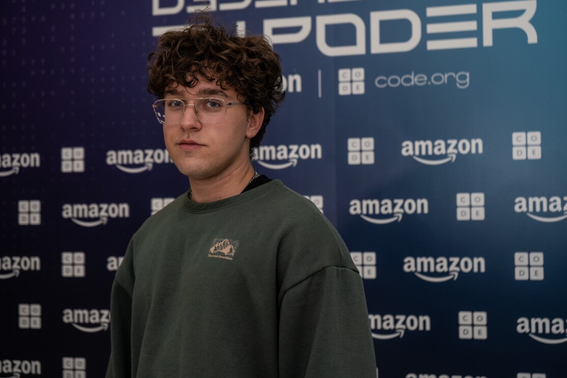 Con un fondo de photocall con el logo de Amazon y de Code.org, en blanco sobre un fondo azul aparece unos de los finalistas, José Jiménez Pajuelo. Lleva  gafas y tiene el pelo rizado. La parte de arriba la lleva más larga. Lleva una sudadera de color verde.