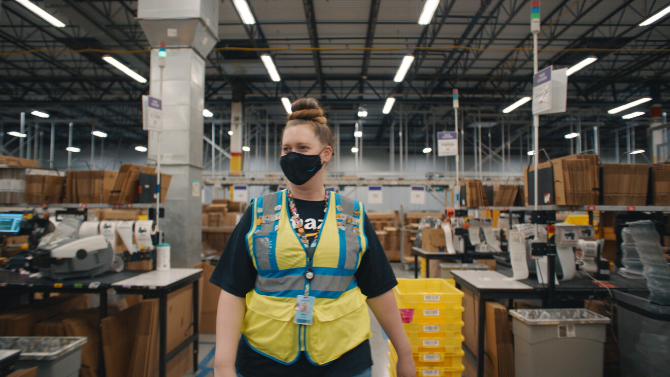 An image of a woman walking through a walkway at Amazon. She is wearing a black mask with the Amazon logo on it and a yellow safety vest. She also had a lanyard with her employee badge on it and the lanyard has many Amazon-related pins on it.