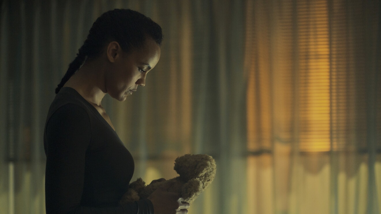 Still from 'Neagley' of woman in black holding a small teddy bear, looking down pensively in dim lighting
