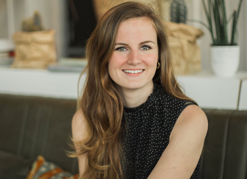 An image of a woman smiling for a headshot photo while sitting on a bench indoors.