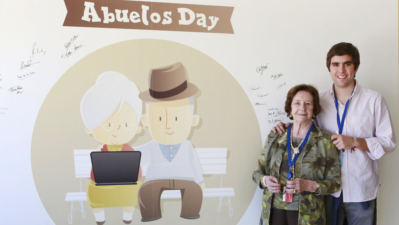 Pedro Armas posa con su abuela al final del tour y delante del cuadro que todos los abuelos han querido firmar como recuerdo. Pedro va vestido sport y su abuela lleva un conjunto de pantalon, camisa y americana.