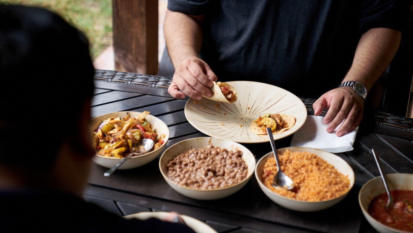 Una mesa con un plato principal en el que come un hombre que solo se le ven las manos y tiene un reloj en el brazo izquierdo y va con un polo azul oscuro de manga corta. Alrededor del plato principal hay tres platos con relleno para las fajitas. El hombre está con una fajita en la mano derecha. Un hombre desenfocado aparece en el margen interior izquierdo de la foto.