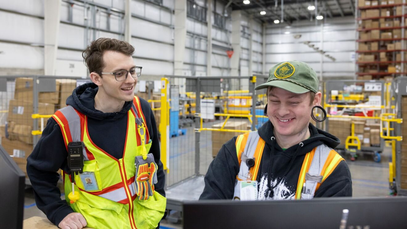 Warehouse workers in safety vests smiling at computer screen.