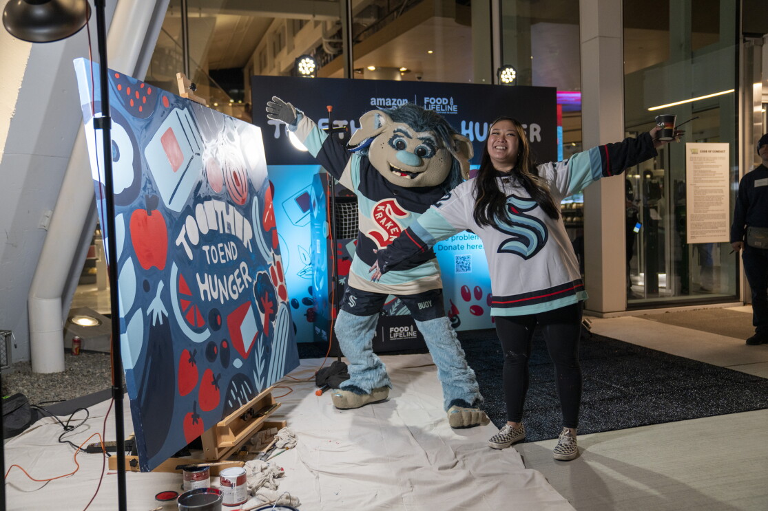 Artist Stevie Shao and Seattle Kraken mascot, Buoy, pose and smile in front of the mural Stevie painted live during the Together to End Hunger food drive.