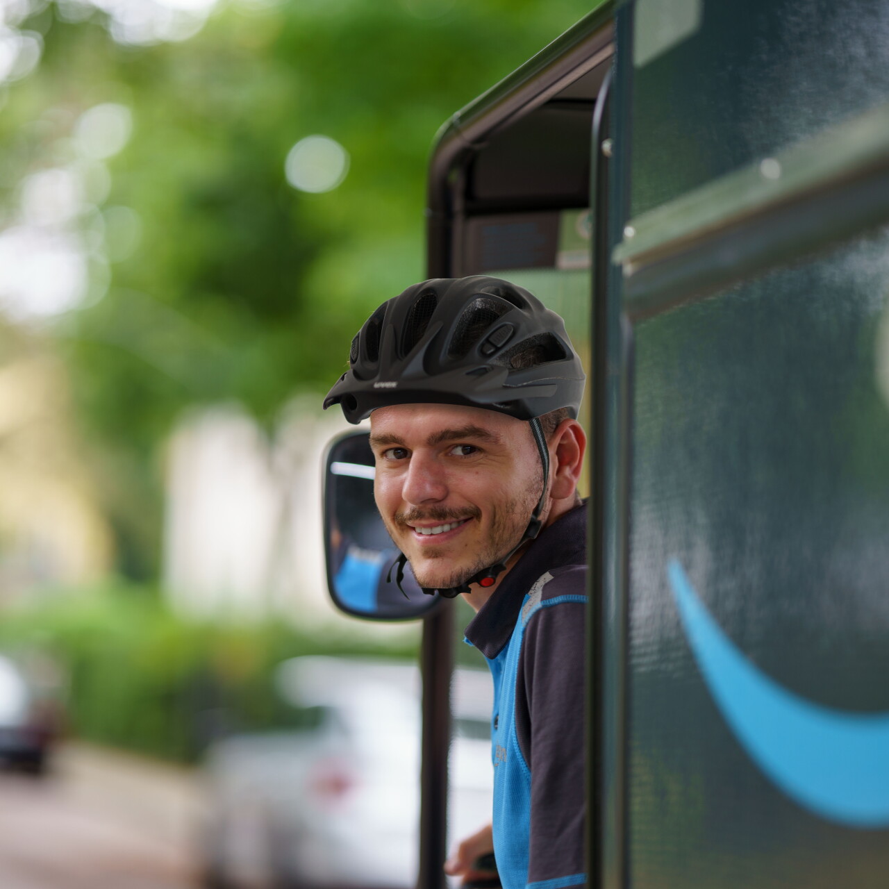 Ein junger Amazon E-Cargo-Bike-Fahrer mit schwarzem Helm und blauem Shirt auf dem E-Cargo-Bike. Er blickt sich um und lächelt in die Kamera. Verschwommen sieht man das seitlich angebrachte Amazon-Logo.