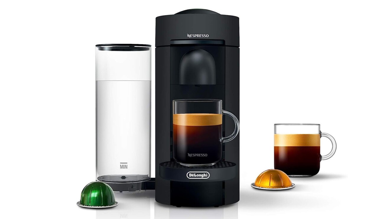 An image of a home espresso machine. The base is black with an adjacent water tank that is clear. There is a clear mug with espresso being poured into it from the machine. Next to the machine are cups of Nespresso refills and another clear cup of espresso.