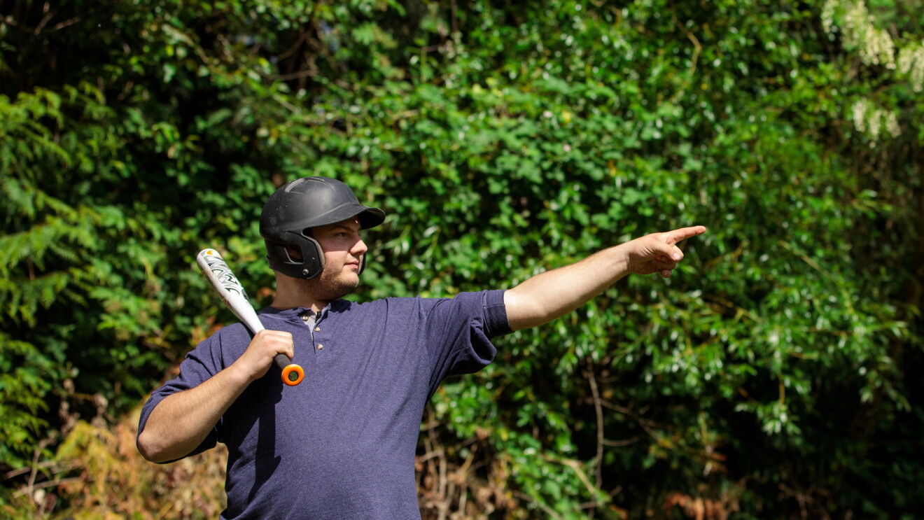 A man balances a baseball bat on his shoulder. He wears a batting helmet. With the hand that's not holding the bat, he points up and away toward the spot he plans to hit the ball.