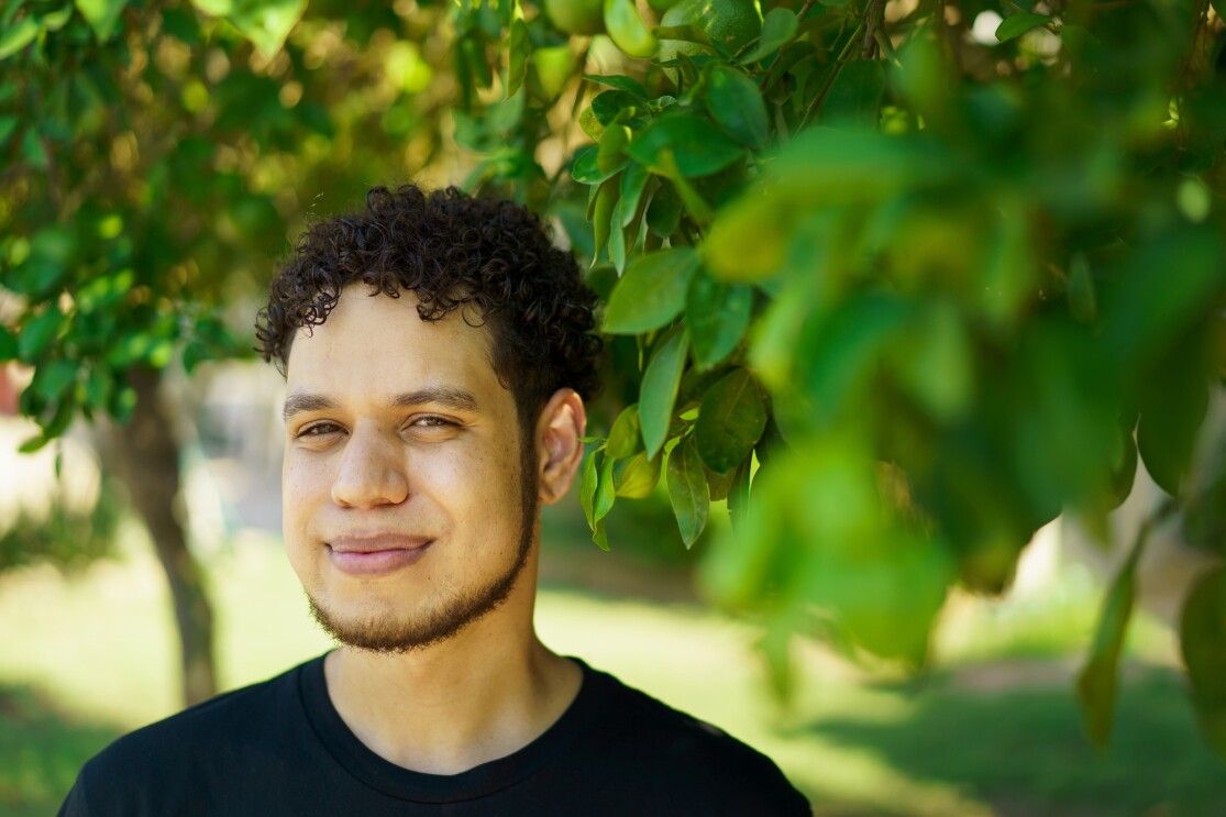 A man in a dark T-shirt is photographed from the shoulders up. He is standing outdoors. Foliage is in the foreground and background.