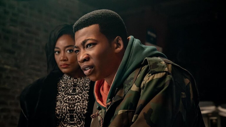 Man in the foreground looking intensely to the side, and a woman standing in the background looking forward with a serious expression