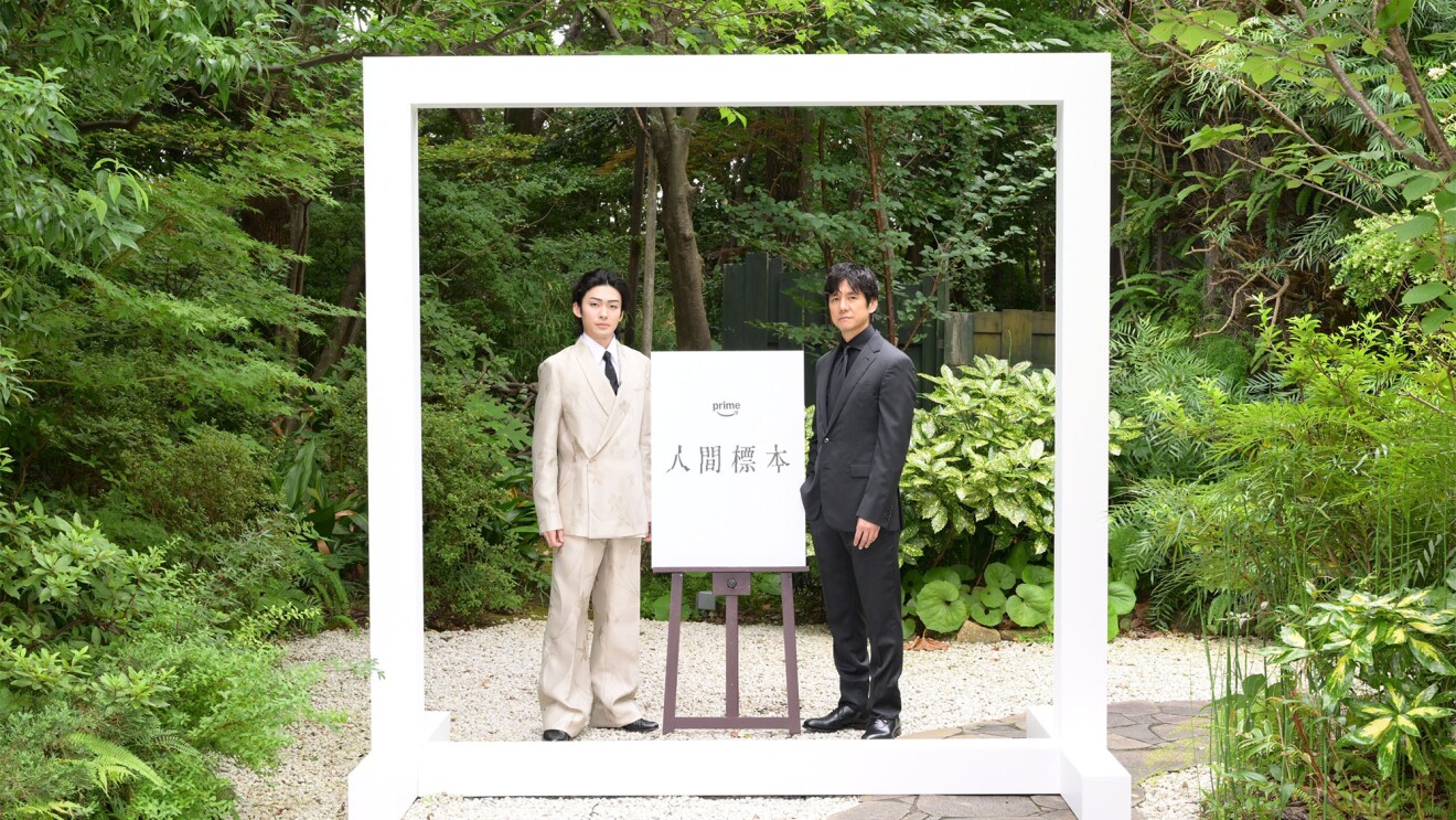 植物が茂る庭にたてられた白いフレームの中に白いスーツを着た男性と黒いスーツを着た男性が立っている。二人の間には「人間標本」と書かれたパネルがイーゼルに立てかけられている