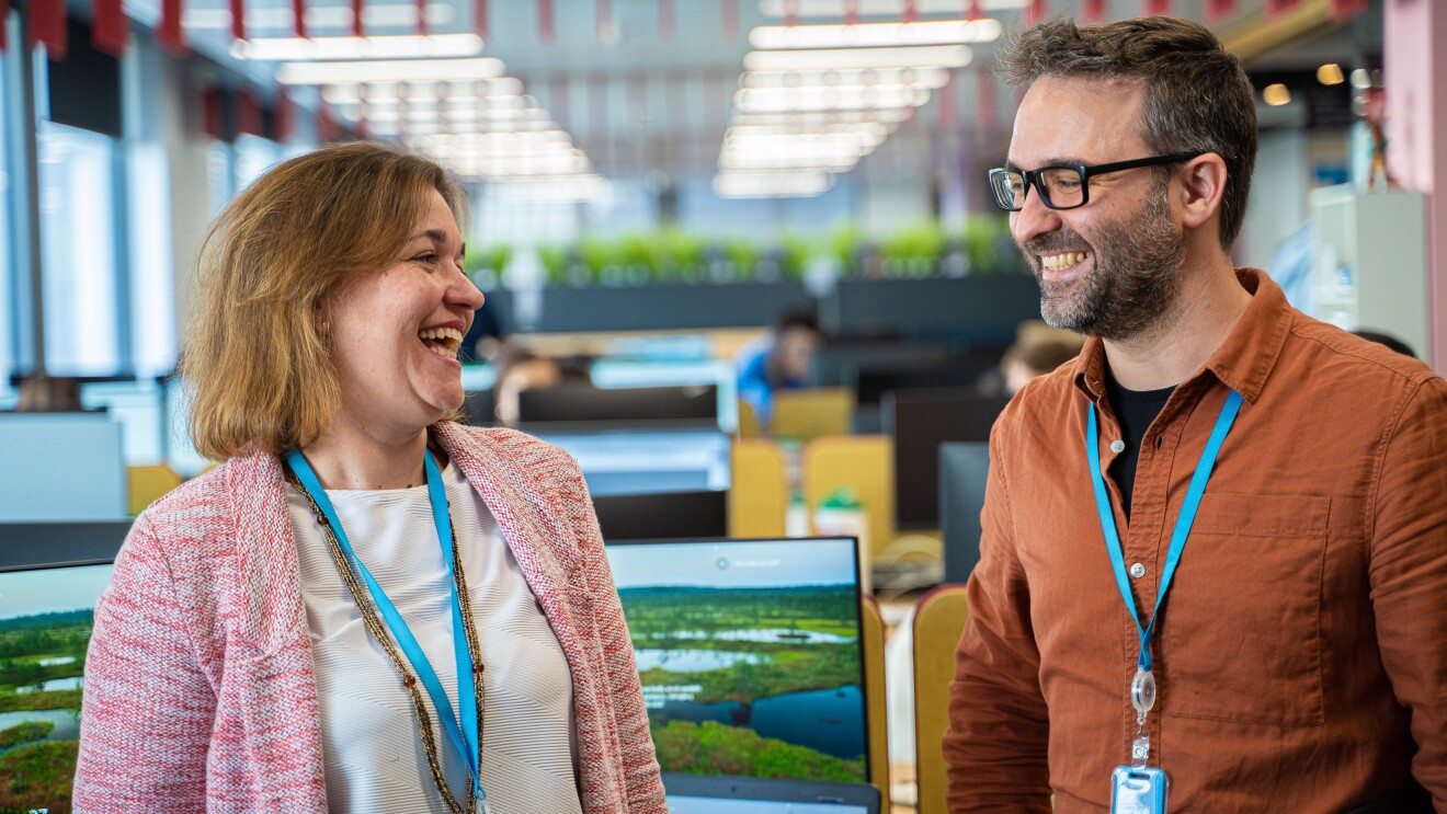 Marta Fernández y Daniel Méndez son Content Program Manager de Alexa. Los dos se miran y sonríen. Marta lleva una camiseta blanca y una chaqueta rosa. Tiene el pelo corto, media melena y es rubia. Daniel lleva una camiseta negra y una camisa marrón. 