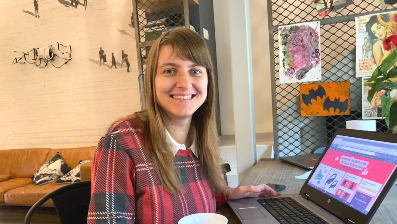 Una donna seduta ad una scrivania davanti ad un pc. Indossa un maglione con fantasia scozzese e in mano regge una tazza nera. Sullo sfondo si intravede un salotto ed una scala. 