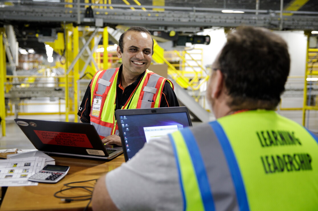 A man working at a laptop smiles at another man working at his own laptop. The two appear to be in conversation.