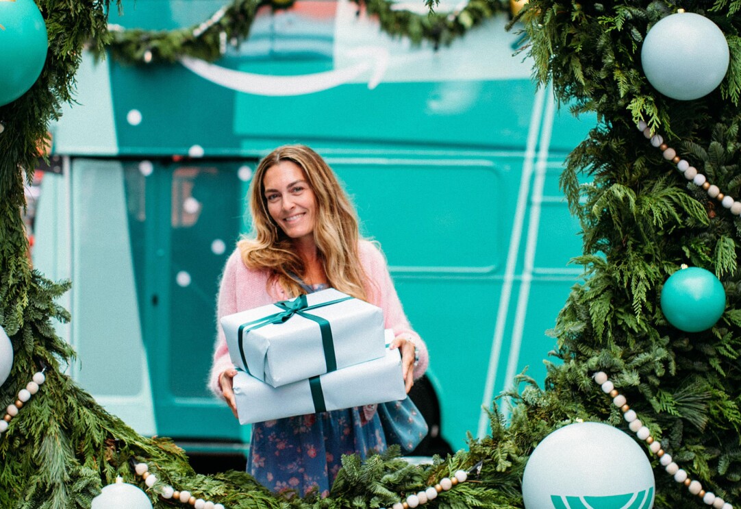 An image of a woman holding two gifts smiling for a photo behind a large wreath.