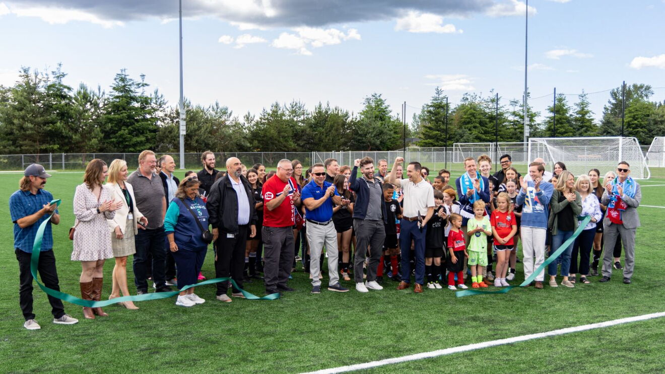 Ribbon cutting celebration with adults and children in soccer uniforms on artificial turf field