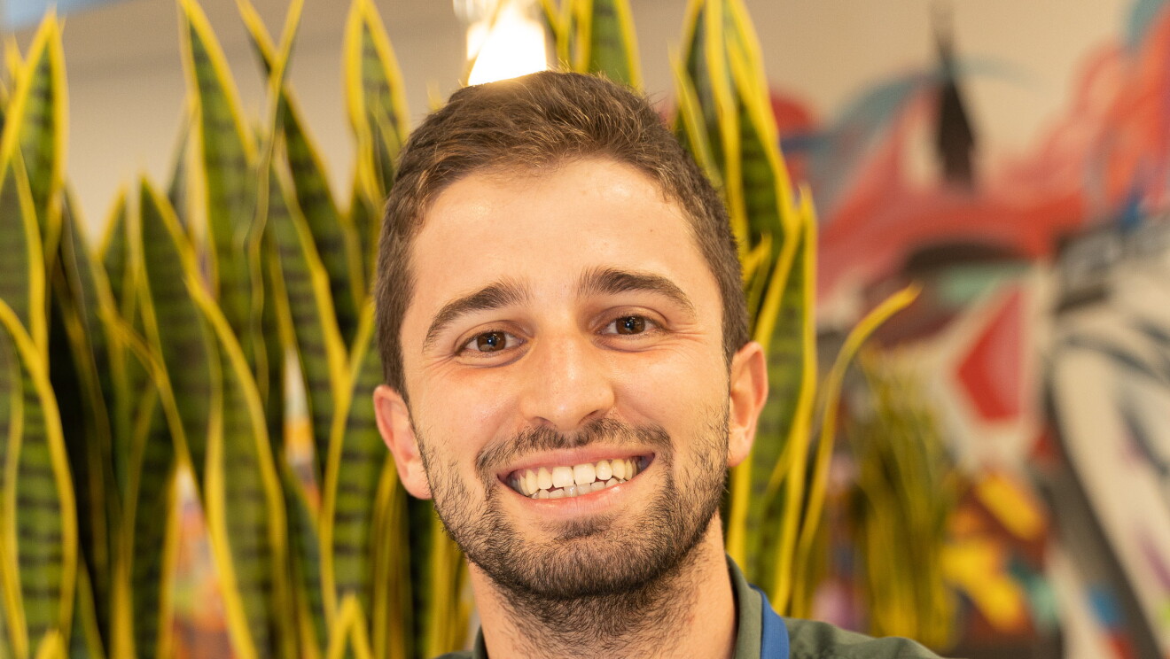 Enrique Fernández, New Account Manager, Channels Sales, mirando a cámara y sonriendo. Lleva una camisa color verde camuflaje y un reloj en la mano izquierda. Sus manos están apoyadas en una mesa. De fondo hay unas plantas de color amarillo y verde y una pared con dibujos de muchos colores. 