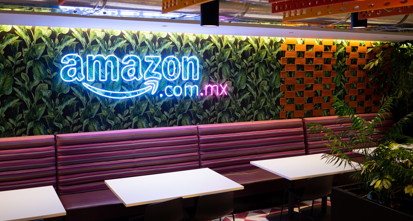 Foto de las oficinas de Amazon. EL techo es visto, con los conductos metálicos y unos plafones de color naranja y rojo. La pared es un jardín vertical y con luces de neón aparece Amazon.com.mx. Con unas mesas de color blanco y unos bancos de color morado.