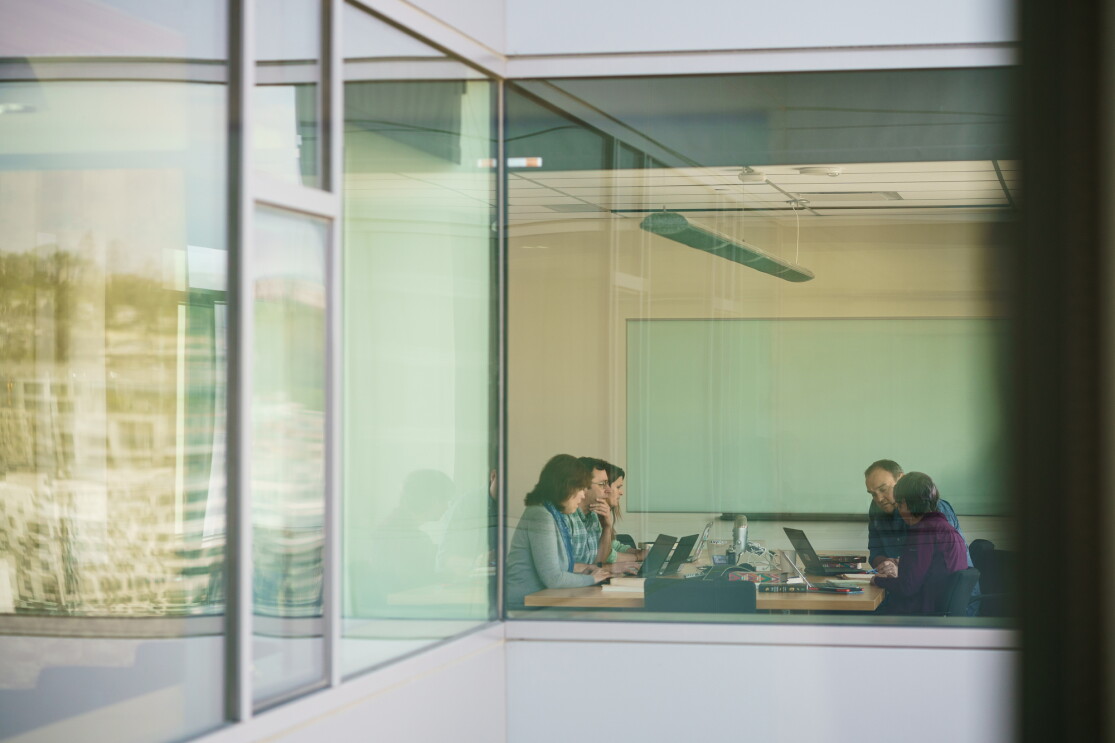 In an image made from another part of the building and photographed through a window, three women and two men sit at a conference table.