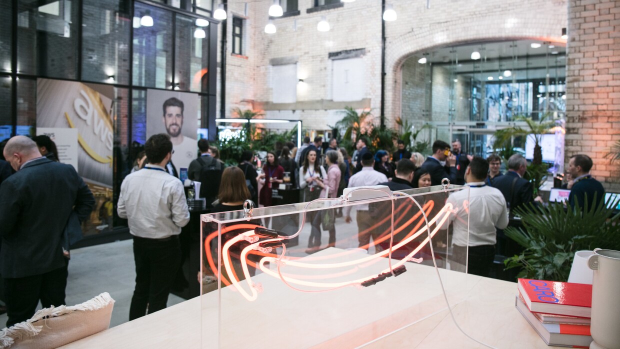A photo of Amazon employees standing and chatting at the Manchester Corporate Office opening. On a table, there is a large Amazon smile logo.