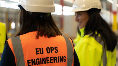 Sonia Corvera, Responsable Senior de Construcción para Amazon España, e Isabel Bermúdez, Responsable Senior de Proyectos de Ingeniería para Amazon España. Las dos protagonistas están de espaldas y llevan casco. Sonia va con un chaleco amarillo e Isabel naranja. Se están mirando y Sonia está sonriendo.
