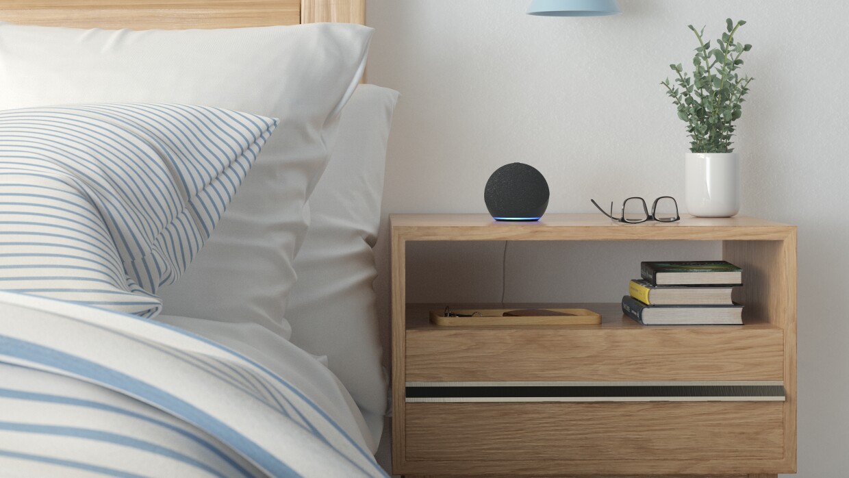 Una habitación con una  cama y una mesita de noche. Los muebles son de madera, estilo pino. El cabecero es alto y la mesita de noche tiene dos cajones. En la cama dos cojines blancos y uno de rayas azules y blancas. Hay un plumón también con el mismo estampado. En la mesita de noche hay un cajón abierto con libros y otro cerrado. Encima de la mesita un Echo Dot de Amazon con la luz azul encenddia, una gafas, una planta verde y con un jarrón blanco. Y una lámpara de pared de color azul.