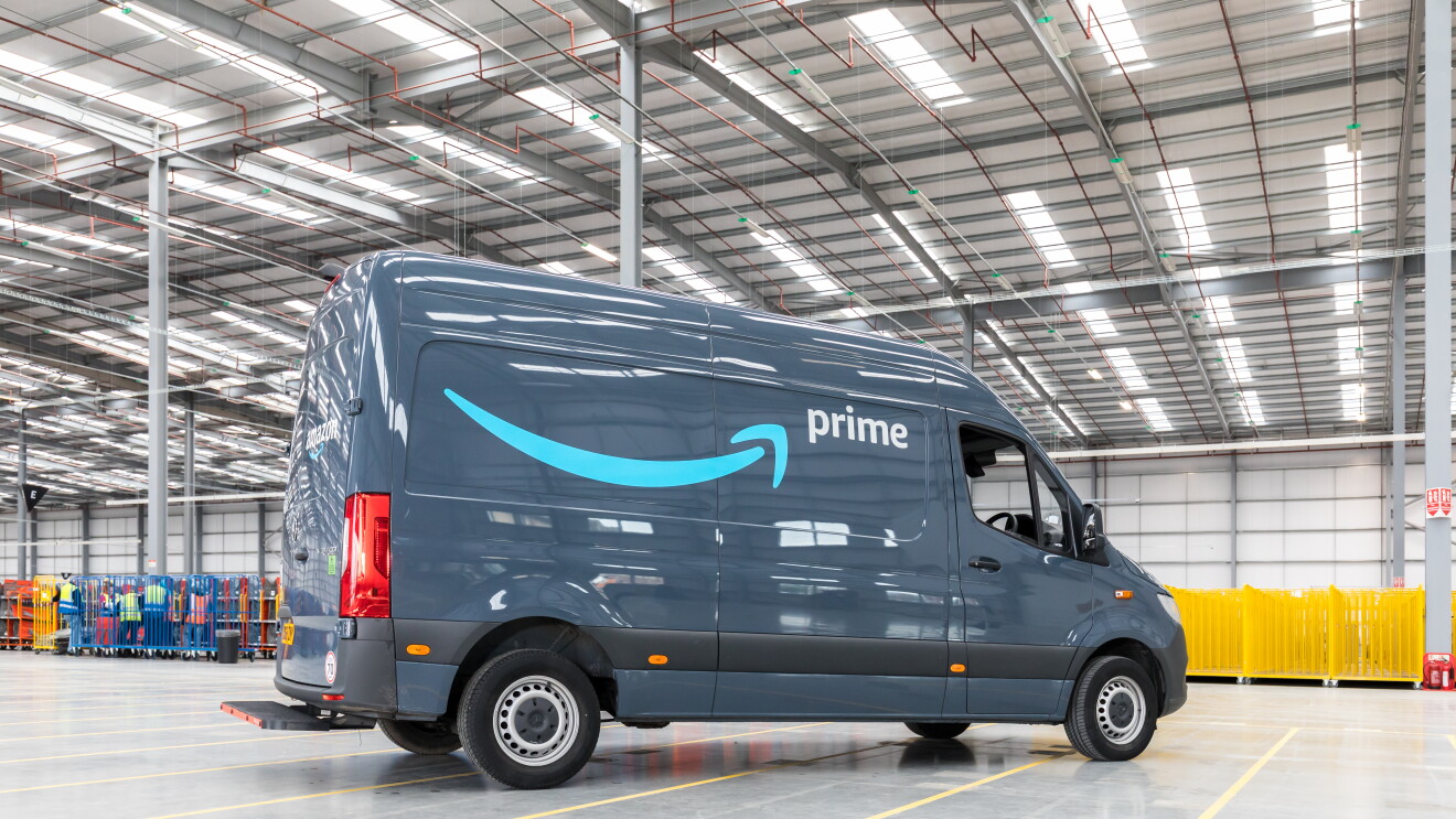 Delivery Service Partner (DSP): la nueva iniciativa de Amazon ayuda a que sus empleados inicien su propia compañía de reparto. Dentro de un centro logístico aparece una furgoneta de color gris con el logo de la sonrisa en azul y la palabra Prime en blanco. 