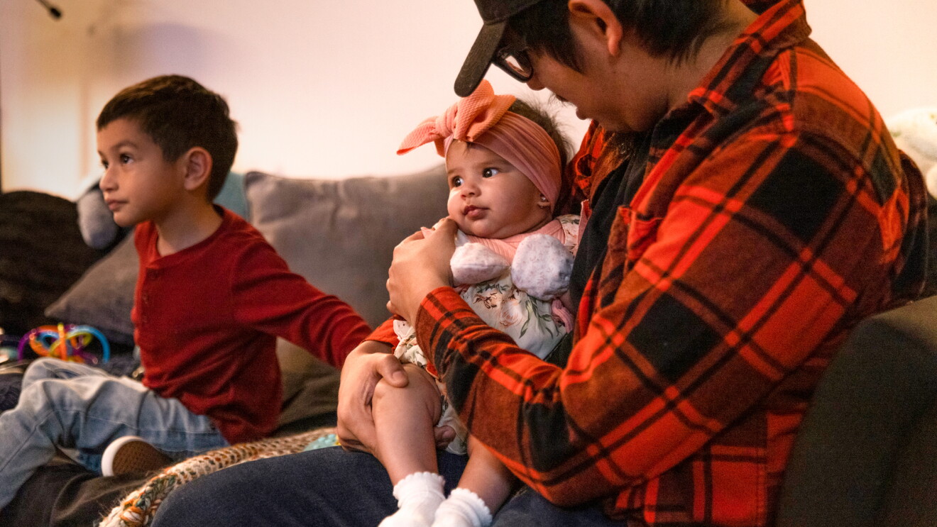 A father sits on a couch holding his newborn daugher and his son sits next to them.