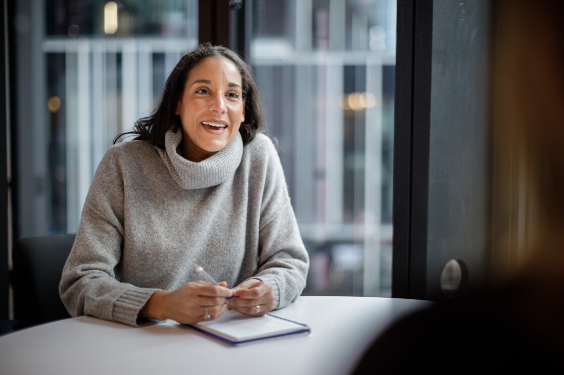 A woman in a grey roll neck sweater speaks to the camera