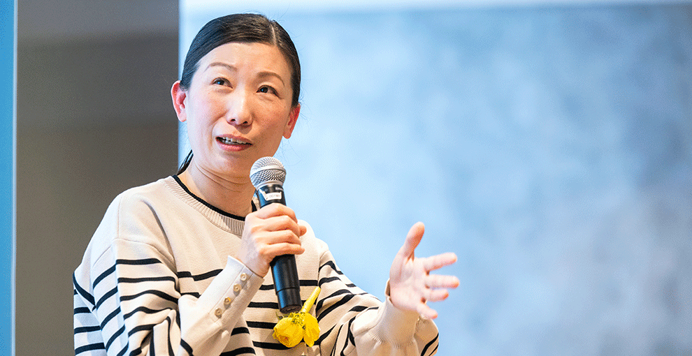 マイクを右手で持った女性。女性の背後にはグレーの壁。