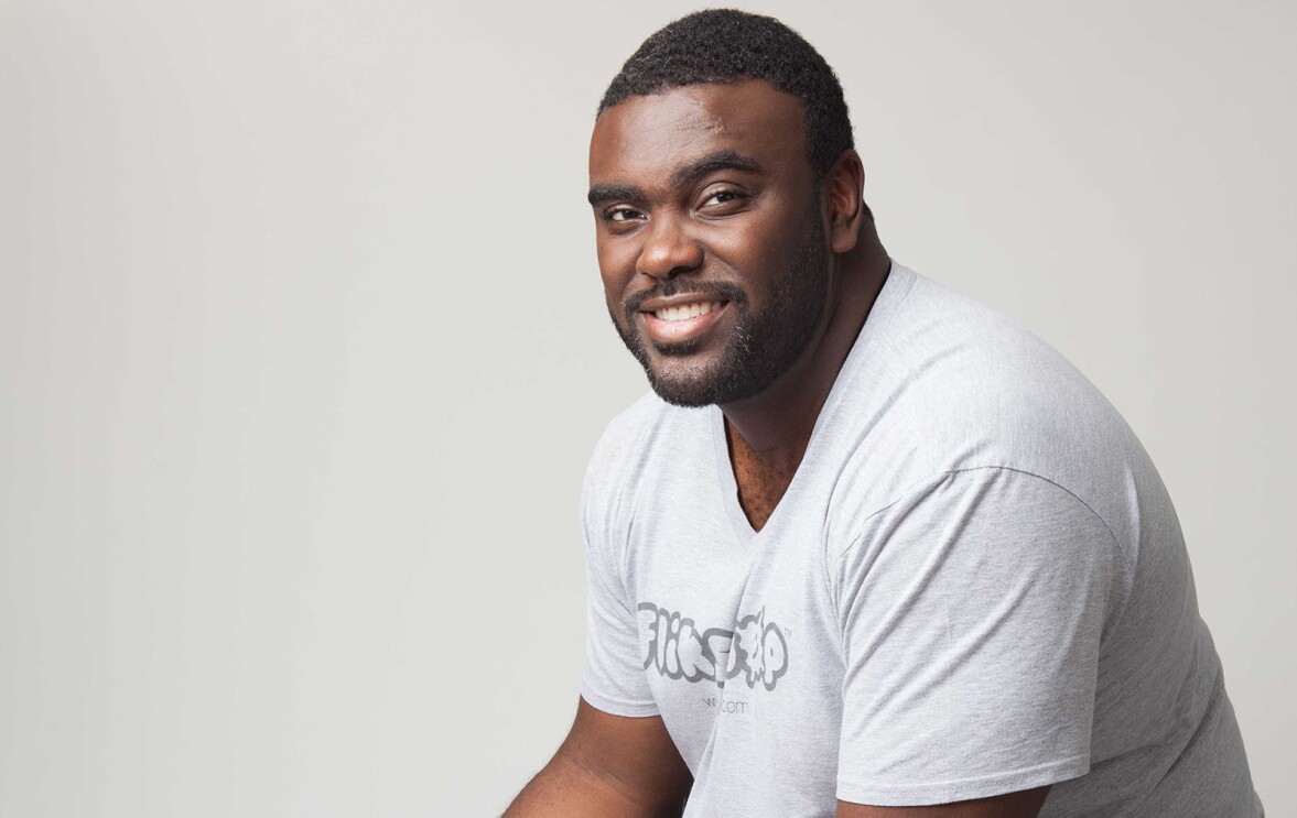 An image of a man smiling for a photo in front of a white background. His shirt says "Flikshop."