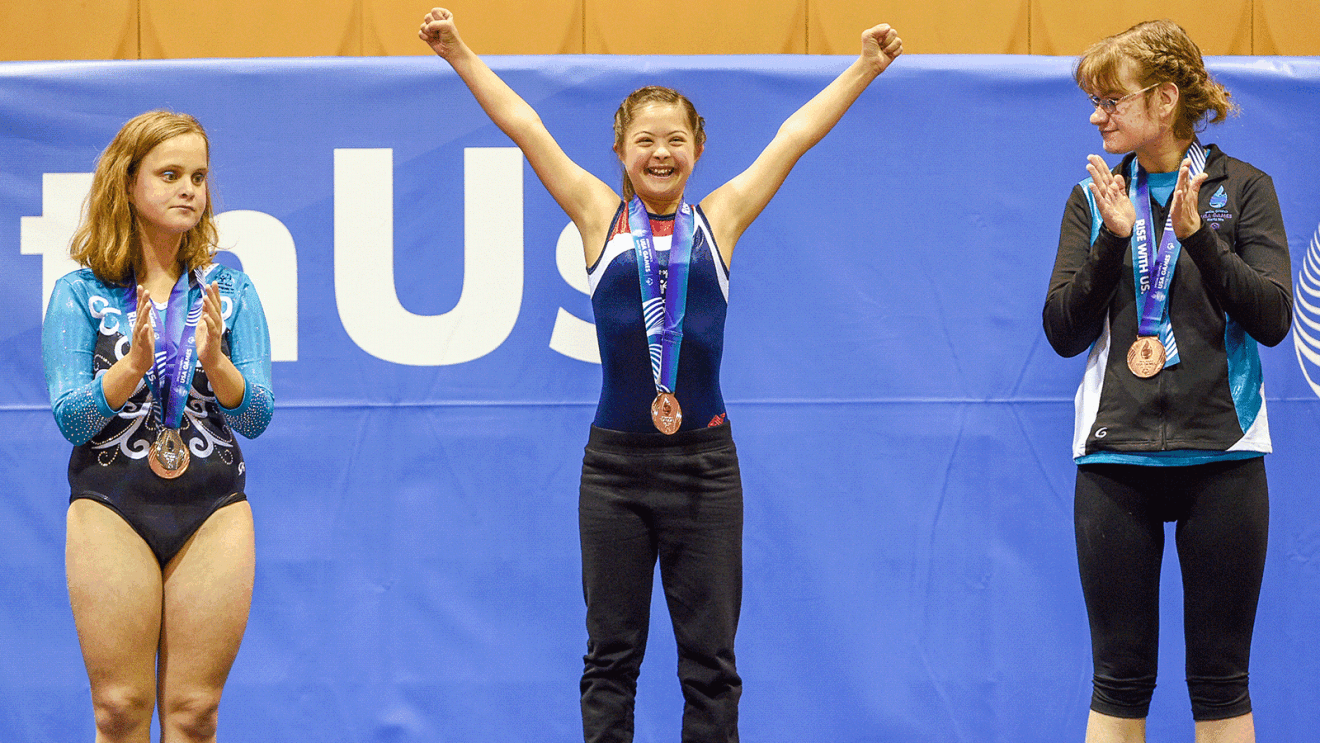 An image of three athletes standing in a podium. The center of the image includes a girl with her arms up wearing a medal. There are two girls, one on each side, wearing medals and clapping. 