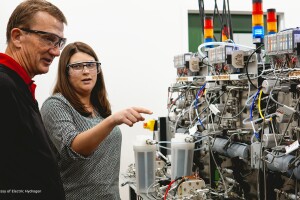 Two people wear safety glasses as they look at the inner workings of a machine.