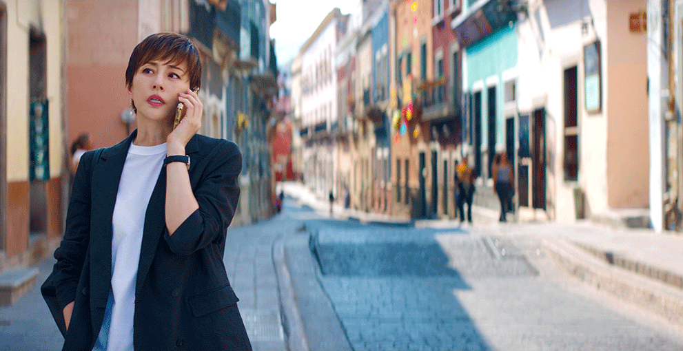 欧米のカラフルな街並みを歩きながらスマートフォンで通話する女性。ショートカットで、黒いジャケット、白い丸襟シャツを着用。