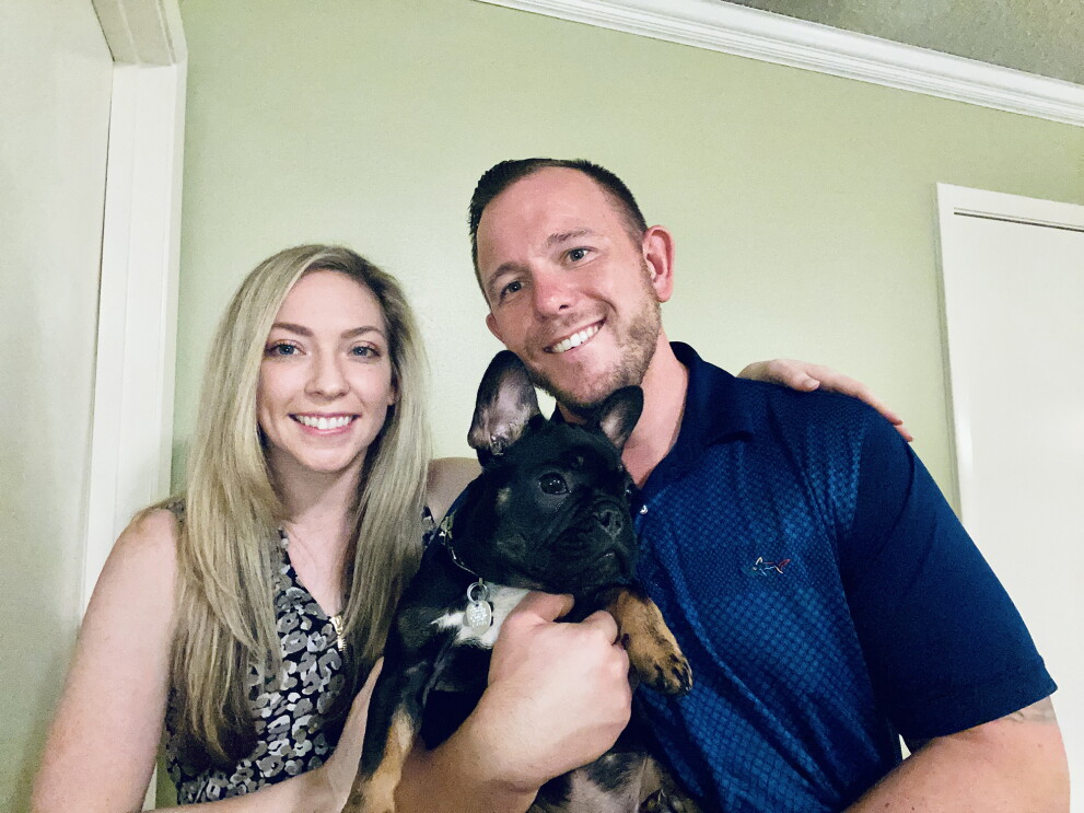 A man and a woman smile for a photo while holding a black French bulldog.