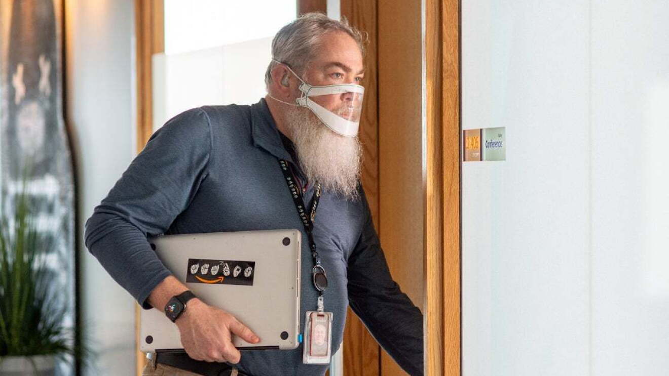 A man wearing a clear face mask carries his laptop computer as he enters a conference room at Amazon's corporate headquarters in Seattle.