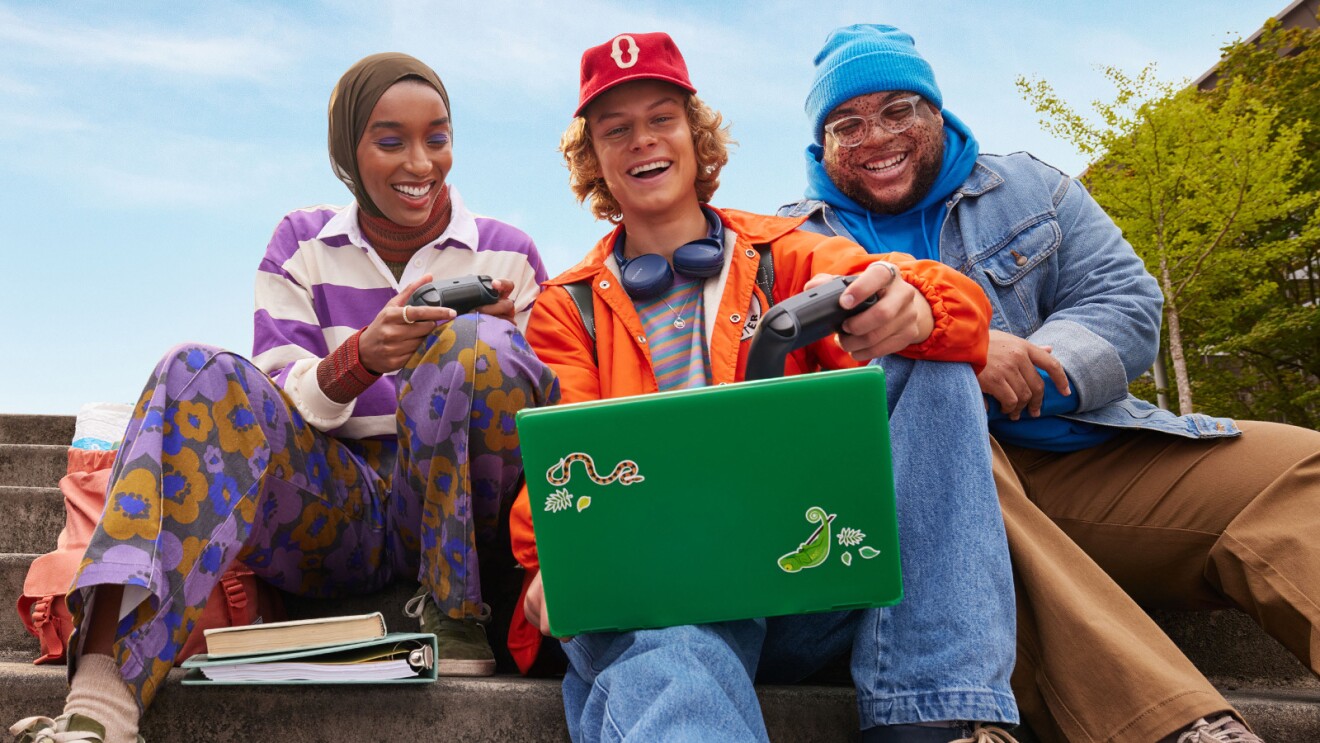 ein Trio bunt gekleideter Schüler:innen, die nebeneinander sitzen, die Controller eines Videospiels halten und ein Spiel auf einem Laptop spielen. 